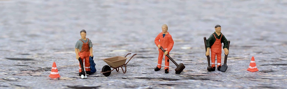 Vahaukot joilla on huomiovaatetus, kottikärryt, lapio, harja ja leka, kaksi kartiota työmaan kumminkin puolin